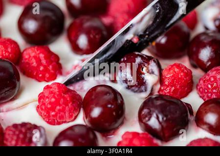Gros plan d'un couteau coupant un cheesecake fait maison garni de cerises et framboises fraîches. Délicieux dessert estival aux baies avec une garniture crémeuse, sélection Banque D'Images