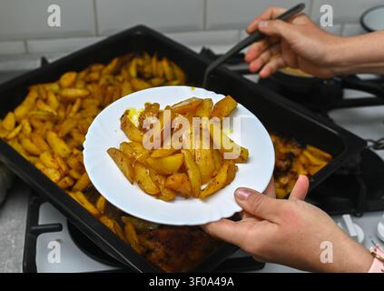 Femme sert des pommes de terre chaudes du plat de cuisson pour le repas de famille. La femme utilise une spatule noire pour déplacer les légumes rôtis. Viande de poulet et morceaux de pommes de terre dorés Banque D'Images