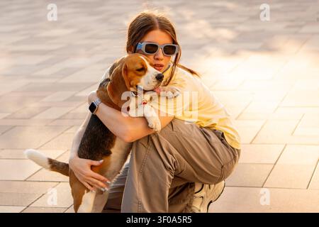 Jeune femme accroupie et câlinant son beagle heureux sous la lumière dorée et chaude du soir. Portrait de style de vie en extérieur mettant en avant la confiance, l'affection et la joie Banque D'Images