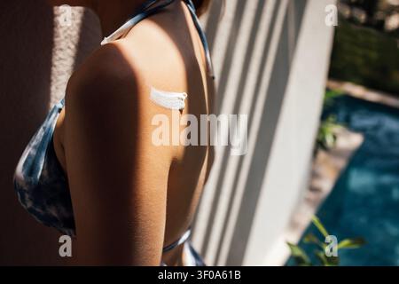 Femme en bikini bleu tie-dye avec un bandage à l'épaule debout près d'une piscine, avec une végétation luxuriante et la lumière du soleil créant une atmosphère vibrante Banque D'Images