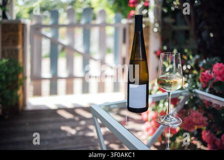 Bouteille et verre de vin blanc posés sur une table dans un jardin, avec une porte en bois et des fleurs colorées en arrière-plan Banque D'Images