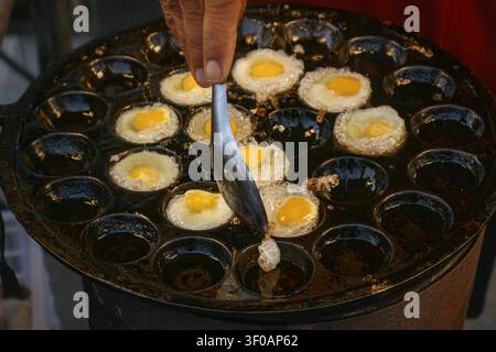Close-up d'œufs de caille frits cuisant sur une poêle traditionnelle en fonte de rue dans un marché thaïlandais avec un vendeur préparant des encas locaux frais. Banque D'Images