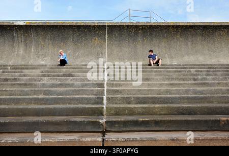 Deux jeunes adultes assis à distance les uns des autres sur les marches d'un mur de défense contre les inondations Banque D'Images