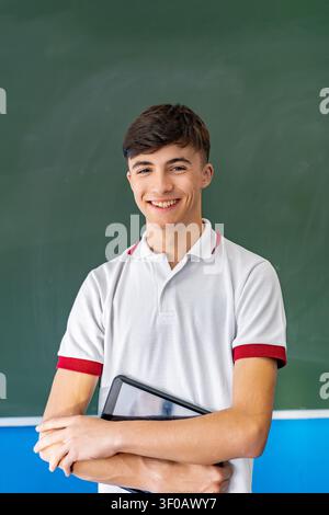 Jeune étudiant debout devant un tableau vert, tenant une tablette et souriant, représentant l'éducation et l'apprentissage Banque D'Images