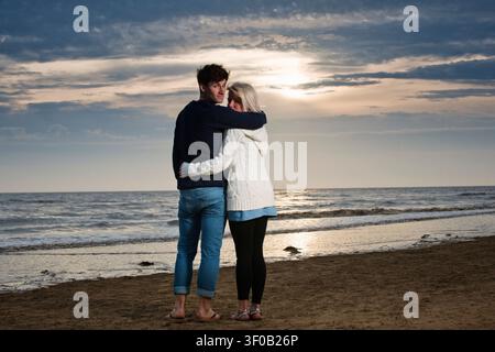 Jeune couple enlacé sur la plage au coucher du soleil regardant vers l'appareil photo Banque D'Images
