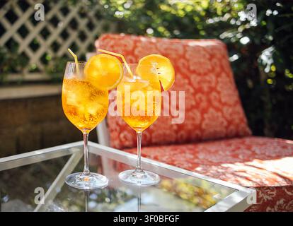 Deux cocktails d'orange rafraîchissants avec glace et tranches de citron reposent sur une table en verre à côté d'une chaise à motifs dans un espace extérieur lumineux du jardin Banque D'Images