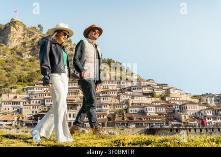 Couple de voyageurs heureux marchant dans la ville historique de Berat sur les collines de l'Albanie avec des maisons ottomanes traditionnelles lors de vacances d'automne ensoleillées et de visite touristique Banque D'Images