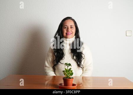 La femme sourit chaleureusement à l'appareil. Elle porte un pull à col roulé crème douillet. Une petite plante en pot est posée sur la table. La plante a des feuilles vertes et charnues. La ba Banque D'Images