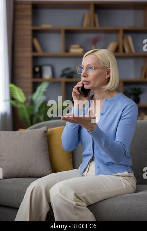 Femme blonde mature portant des lunettes et un cardigan bleu, parlant au téléphone portable tout en étant assise sur un canapé confortable dans un salon lumineux, gesticulant d'une main pendant la conversation Banque D'Images
