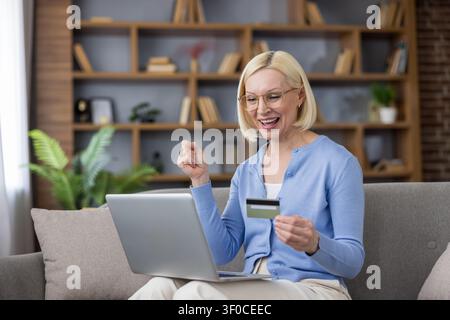Sénior célébrant avec enthousiasme son succès en effectuant un achat en ligne, utilisant un ordinateur portable et tenant une carte de crédit sur un canapé à la maison Banque D'Images