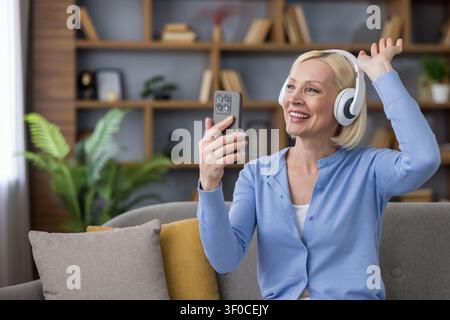 Sénior souriante portant des écouteurs, tenant un smartphone et faisant un signe de la main pendant un appel vidéo, communiquant avec la famille ou des amis tout en se relaxant sur un canapé à la maison Banque D'Images