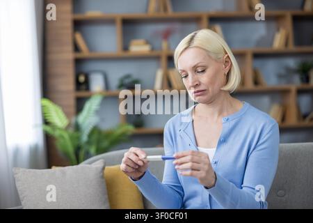 Femme mûre assise sur un canapé chez elle, regardant vers le bas avec une expression sombre tout en tenant un test de grossesse négatif, indiquant des préoccupations liées à la ménopause ou à l'infertilité Banque D'Images