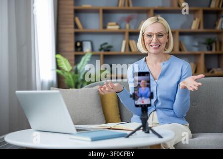 Sénior créant du contenu en ligne et enregistrant un appel vidéo avec un smartphone sur un trépied tout en étant assise sur un canapé à la maison, offrant un enseignement à distance et un apprentissage en ligne Banque D'Images