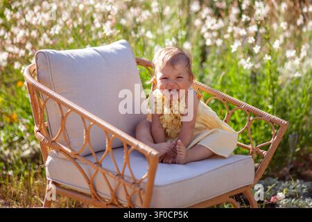Portrait de jeune fille assise sur une chaise dans le jardin d'été. Enfant heureux riant se détend à l'extérieur parmi les fleurs en fleurs Banque D'Images