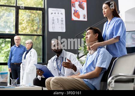 Jeune patient adulte portant une minerve examiné par une infirmière tandis qu'un médecin donne des nouvelles de la guérison. Une assistante médicale féminine ajuste un col cervical sur un homme asiatique, en assurant un soutien et des soins appropriés. Banque D'Images