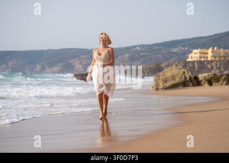 Femme aux cheveux blonds courts portant une robe en dentelle blanche, marchant pieds nus le long du rivage de l'océan sur une plage de sable. Banque D'Images