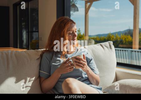 Une femme rousse se détend sur un canapé confortable, tenant une tasse chaude et profitant d'une lumière douce du matin à travers de grandes fenêtres donnant sur une terrasse et Banque D'Images