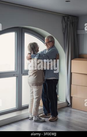 Couple de personnes âgées assis par terre parmi des cartons de déménagement, souriants et utilisant une tablette tout en se détendant, planifiant et déballant en s'installant dans leur nouvelle maison ensemble Banque D'Images