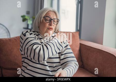 Femme âgée sur un canapé de salon serrant son épaule avec une expression douloureuse, exprimant l'inconfort, le vieillissement et les préoccupations de santé dans un cadre domestique Banque D'Images