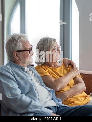 Un couple âgé est assis près l'un de l'autre sur un canapé avec le bras du mari autour de sa femme, souriant et profitant d'un moment calme et affectueux à la maison, transmettant lo Banque D'Images