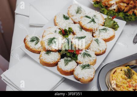 Tranches de pain grillé dorées garnies d'herbes fraîches et de fromage râpé. Présentées avec élégance avec des verts vibrants et des poivrons rouges. Servies sur une table blanche impeccable Banque D'Images