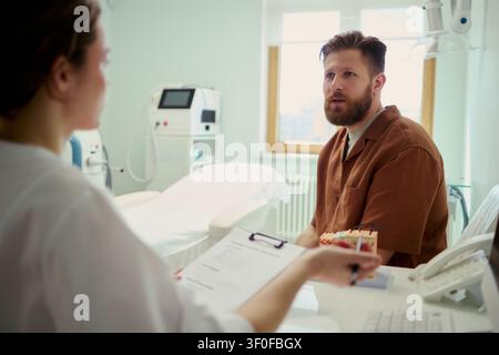Jeune homme caucasien consultant avec une clinicienne dans un cabinet médical, discutant du plan de traitement et de soins. Utile pour le marketing sanitaire, la communication avec les patients, les services de clinique Banque D'Images