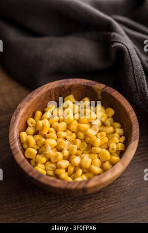 Un gros plan de grains de maïs doux jaunes vifs servis dans un bol en bois artisanal sur une table en bois rustique avec une nappe en lin sombre, évoquant la chaleur Banque D'Images