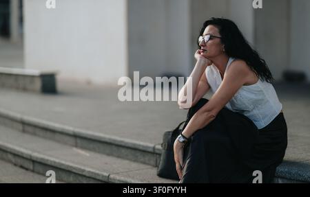 Femme pensive assise sur des marches extérieures en ville, portant des lunettes et une tenue d'été décontractée Banque D'Images