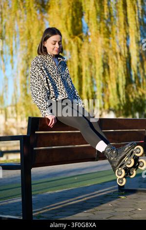 Jeune patineur profitant d'un moment de calme en plein air, assis sur un banc avec les jambes étendues sous une lumière chaude du jour. Idéal pour le bien-être, la détente, les loisirs urbains ou la publicité saisonnière Banque D'Images