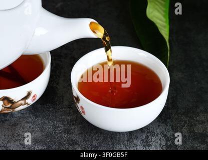 Verser du thé chaud d'une théière en céramique blanche dans une tasse à thé assortie sur une surface sombre Banque D'Images