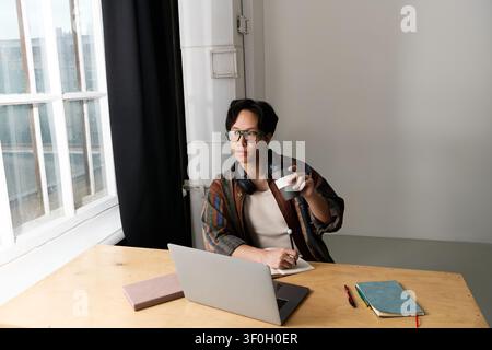 Jeune homme en tenue élégante assis à un bureau en bois, sirotant un café et concentré sur ses tâches. Banque D'Images