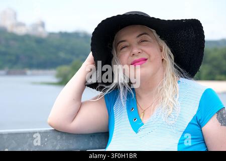 Belle femme mûre souriante de plus de 50 ans portant un chapeau à larges bords noir et une tenue d'été bleue, posant en plein air au bord de l'eau par une chaude journée ensoleillée. Nat Banque D'Images