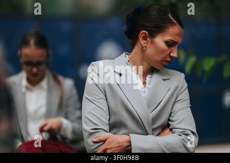 Femme d'affaires en costume gris debout les bras croisés, l'air pensif tandis qu'un collègue est flou derrière elle Banque D'Images