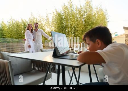 Famille heureuse sur le balcon d'une maison moderne avec un enfant utilisant un ordinateur portable pendant que les parents profitent de la vue sur la mer depuis la terrasse Banque D'Images