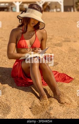Jeune femme cubaine attirante en bikini rouge feuilletant les pages d'un livre assise sur la plage de sable Banque D'Images