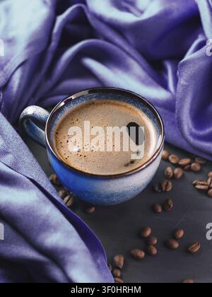Une tasse bleue de café noir fraîchement infusé sur une table sombre avec des grains de café éparpillés Banque D'Images