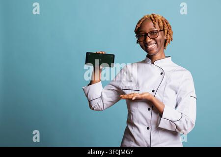 Femme noire en uniforme de chef tenant un smartphone avec écran vert sur fond bleu. Cuisinière professionnelle souriante présente un espace numérique à copier pour des promotions culinaires ou des annonces de restaurant. Banque D'Images