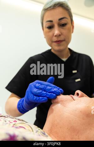 Une femme âgée se détend pendant un traitement professionnel de soins de la peau, tandis qu'une esthéticienne gantée applique de la crème sur son visage dans un cadre de spa clinique propre. Banque D'Images