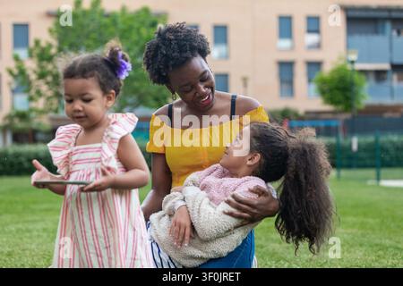 Mère heureuse passant du temps avec ses deux filles dans un parc verdoyant, partageant une câlin chaleureux et un moment ludique ensemble en plein air près d'une construction d'appartements modernes Banque D'Images