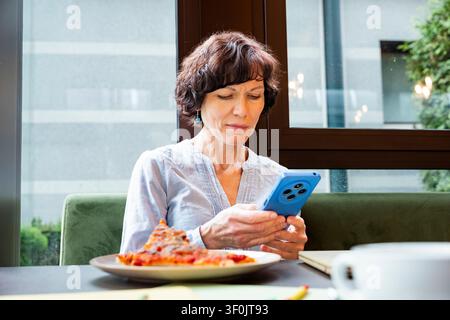 Une femme d'affaires dans un café prend une pause déjeuner, concentrée sur son smartphone. Elle est assise près d'une grande fenêtre, avec une pizza et du café sur la table, dans un m Banque D'Images