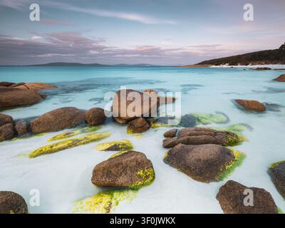 Vue de Whalers Cove avec de gros rochers de granit couverts de mousse reposant dans une eau turquoise peu profonde sous un ciel pastel doux à Albany, WA, Australie. Banque D'Images