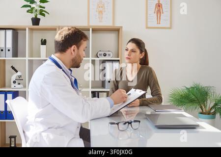 Médecin masculin en uniforme médical blanc consultant une patiente féminine, clinique de consultation, médecin généraliste parlant Banque D'Images