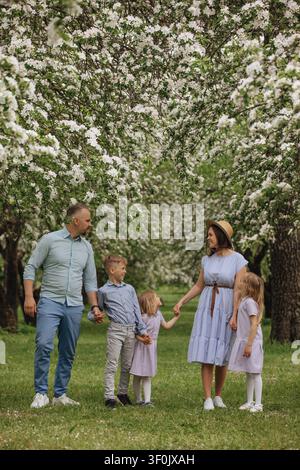 Famille heureuse de cinq personnes marchant main dans la main sous des arbres fruitiers en fleurs dans un verger printanier, profitant d'un moment paisible en plein air sur l'herbe verte avec des blan Banque D'Images