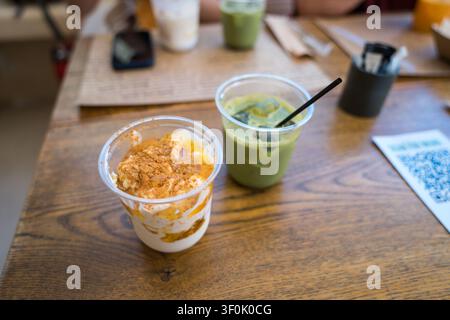 Close-up d'un parfait au yaourt garni de miettes à côté d'un latte glacé au matcha dans des gobelets en plastique sur une table de café en bois, avec des menus et un téléphone flous en arrière-plan Banque D'Images
