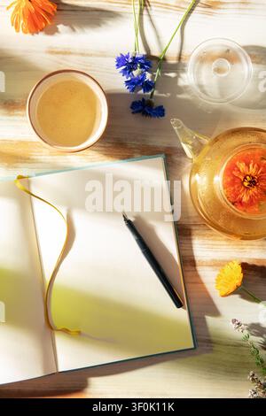 Table en plein air baignée de soleil avec journal ouvert et stylo, thé aux herbes dans une théière en verre et une tasse, et des fleurs fraîches, créant un rituel domestique calme pour une l attentive Banque D'Images