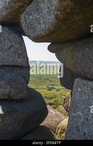Vue à travers la formation rocheuse de Cheesewring sur le Bodmin Moor, révélant un paysage ondulant vert avec des champs sous un ciel bleu clair, Cornouailles. Banque D'Images