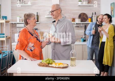 Un couple de personnes âgées se regarde avec des verres de vin dans la cuisine tandis que des jeunes parents prennent des photos avec leur téléphone lors d'une réunion de famille. Banque D'Images
