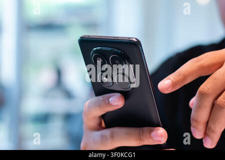 Une image en gros plan montrant une main humaine tenant un appareil mobile noir élégant avec une configuration triple caméra distincte à l'arrière, utilisé à l'intérieur avec une lumière douce Banque D'Images