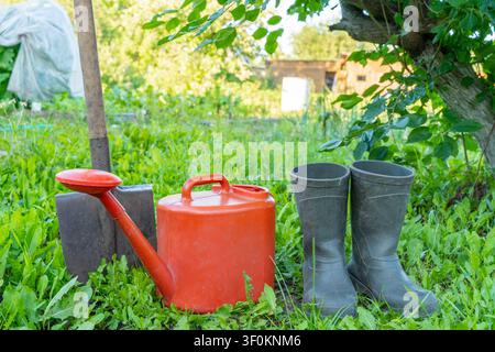 Équipement de jardinage avec arrosoir rouge, bottes et pelle sur l'herbe dans un cadre de jardin luxuriant. Style de vie éco-responsable, agriculture naturelle Banque D'Images