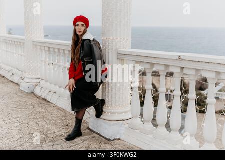 Femme en mode béret posant sur un balcon blanc avec vue sur l'océan côtier et guirlandes lumineuses pendant la saison hivernale Banque D'Images